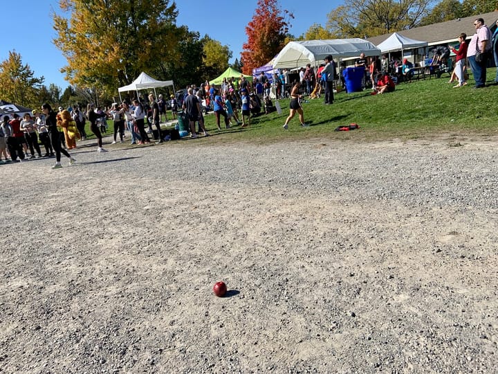 a red apple lying on a gravel trail with tents and people milling about in the distant background on a sunny day under blue skies